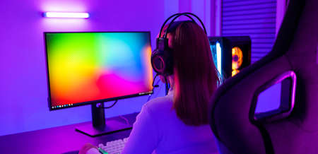 Asian professional gamer playing online video game on desktop computer PC have colorful neon LED lights dark room, young woman in gaming headphones using computer for playing game at home, Back viewの写真素材