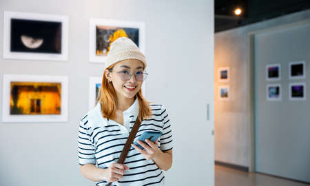 Visitor woman standing takes picture art gallery collection in front framed paintings pictures with mobile phone, Asian female watching at photo frame with smartphone at artwork gallery showの写真素材