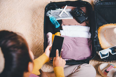 Woman using smartphone blank screen and packing suitcase bag for travel trip weekend vacation at home, female check information in tourism website on mobile phone before go to airportの写真素材