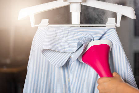 Close up of woman using electric hand steam generator while it is hot and steam comes out for clothes shirt at house, Household & home equipment for housework, concept of linen careの写真素材