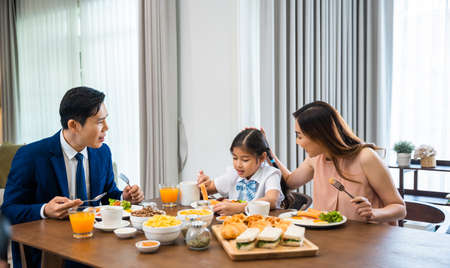Asian family father, mother with children daughter eating healthy breakfast food on dining table kitchen in mornings together at home before father left for work, happy couple adult family conceptの写真素材