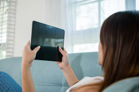 Happy Asian woman playing smart tablet at home while relaxing on sofa living room, Beautiful female sitting on couch using digital tablet pc, technology social mediaの写真素材