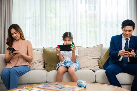 Family don't care about each other. Asian parents ignore their child and looking at their mobile phone at home, father and mother read social media but daughter play video game on sofa living roomの写真素材