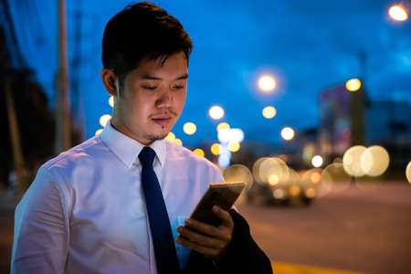 Young man walking and chatting on mobile phone with friends at social networks outdoor, Portrait Asian businessman typing an sms message via smartphone after work near office at night city streetの写真素材