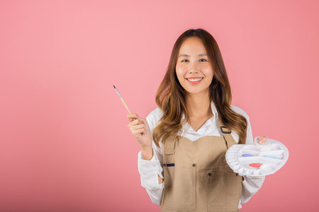 Happy female painting using paintbrush and palette with colors, Asian beautiful young woman artist holding brush and paint palette, studio shot isolated on pink background, Paintings and art equipmentの写真素材
