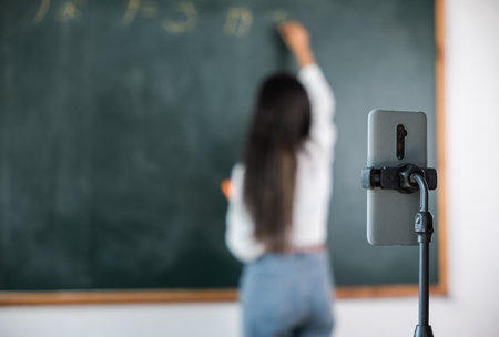 Back to school online class. Asian teacher teach on board while have blackboard lecture online through smartphone in classroom, woman standing front of class live video call online smart mobile phoneの写真素材