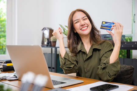 Happy Asian freelancer holding credit card she excited raised hands say yes! do winner gesture on...