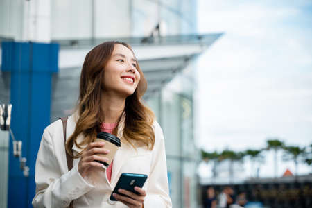 Young business woman smiling holding mobile phone with coffee take away going to work early in...
