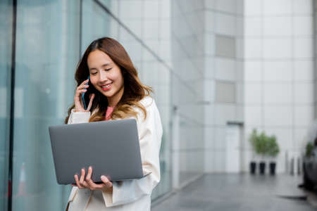 Asian businesswoman working on laptop and talking on cell phone at front building near office, Portrait beautiful business woman in white suit smile discussing issues on smart mobile phone in cityの写真素材