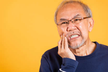 Dental pain. Portrait senior old man sad hand touching cheek suffering from toothache studio shot isolated on yellow background, Asian unhappy elder man problems with teeth pain, dental healthcareの写真素材