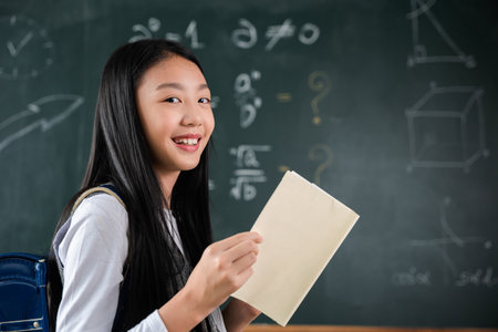 Back to school. Happy beautiful Asian Schoolgirl girl standing holding books standing in front blackboard of classroom, Portrait of smiling woman child student of black chalk board, Education lessonの写真素材