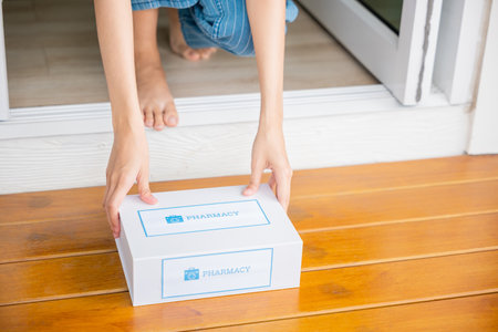Close up hands of sick Asian woman sitting at door to receive medication first aid pharmacy box from hospital delivery service at floor home, merchandise medicine online business, healthcare conceptの写真素材
