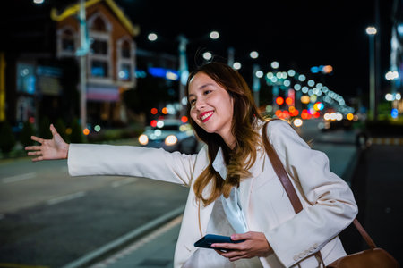 Beautiful woman smiling using smartphone application hailing with hand up calling cab after late work, Asian businesswoman standing hail waving hand taxi on road in busy city street at nightの写真素材