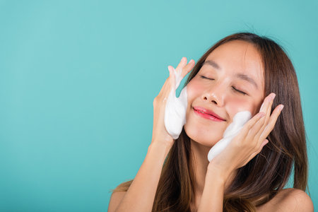 Asian woman face wash exfoliation scrub soap with skincare cleansing, Portrait beautiful female smile applies foaming cleanser healthy skin, Studio isolated on blue background, skincare daily routineの写真素材