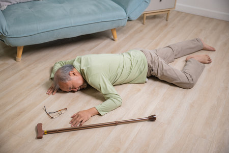 Asian senior man falling down lying on floor at home alone with wooden walking stick in living room, Elderly man headache after fall down, Health care and medicine conceptの写真素材
