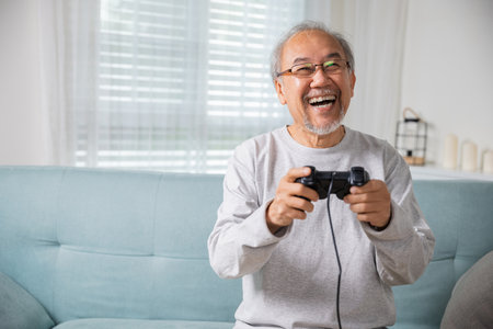 Asian senior old man enjoying holding joystick playing video game at home in living room, mature man hands using game controller, Funny on retirement elderly smile sitting on sofa life gamingの写真素材