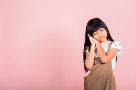 Asian little kid 10 years old standing with hands sealed pretending he is sleeping closed eyes at studio shot isolated on pink background, Happy child girl holding hands under face tired dreamingの写真素材