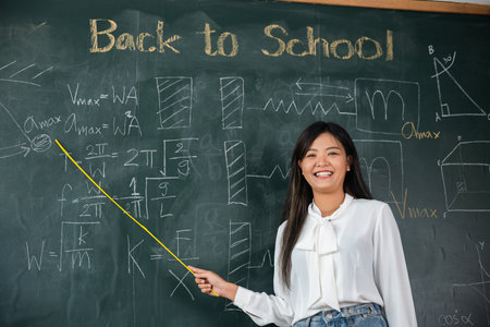 Back to school concept. Happy beautiful young woman standing hold pointer to back board, Asian female teacher smiling with wooden stick pointing to blackboard at school in classroom, Educationの写真素材