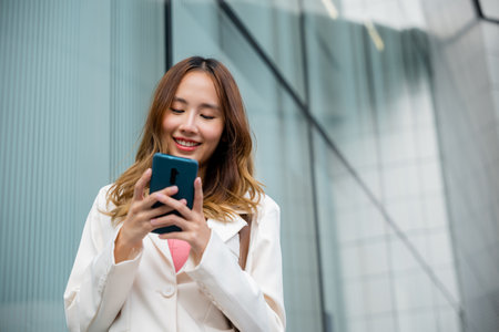 Asian businesswoman texting smartphone commuting work she walking near her office building in morning, Beautiful business woman smiling hold mobile phone outdoor walking on city street urbanの写真素材