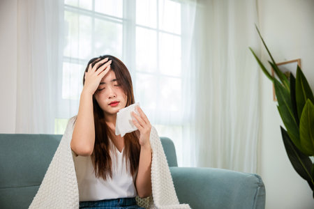 Sick woman sitting under blanket on sofa and sneeze with tissue paper in living room, Asian young female blowing nose sneezing in tissue at home, fever caught coldの写真素材
