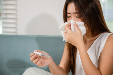 Sick woman have symptom sick high fever she checking her temperature with thermometer in living room at home, young female suffering from flu cold and fever, health care and medical conceptの写真素材