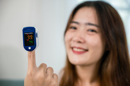 Asian young woman using medical oximeter equipment to measure pulse oxygen saturation on sofa in...