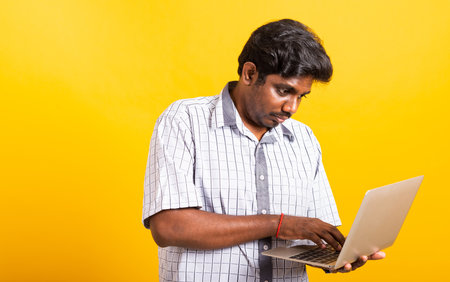 Asian happy portrait young black man smiling standing wear shirt holding and typing laptop computer he looking to camera isolated, studio shot yellow background with copy spaceの写真素材