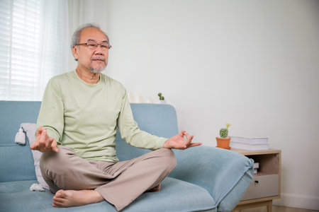 Lifestyle calm senior man lotus pose doing yoga for mental balance breathing air relaxing on sofa at home, Asian elderly old man sitting practise yoga and meditation in lotus position and closed eyesの写真素材