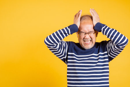 Portrait senior old man sad suffering from migraine studio shot isolated on yellow background, Asian elder man hands holding head and close ears suffering from headache, depressed healthcare, amnesiaの写真素材
