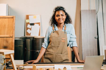Portrait of africa american beautiful young black woman carpenter wearing protective goggles on head standing on workspace table at workshop against wood or woodshop, National Carpenters Dayの写真素材