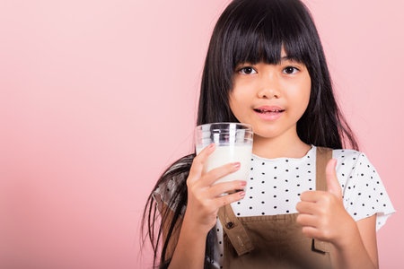 Asian little kid 10 years old smile hold milk glass drink white milk and show thumb up finger for good sign at studio shot isolated on pink background, Happy child girl daily health care medicine foodの写真素材