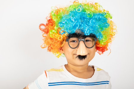 April Fools Day. Portrait of Funny kid boy clown wears a curly wig colorful a big nos and glasses and has a mustache isolated on white background with copy space, Happy child festive decorの写真素材