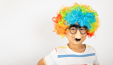 April Fools Day. Portrait of Funny kid boy clown wears a curly wig colorful a big nos and glasses and has a mustache isolated on white background with copy space, Happy child festive decorの写真素材