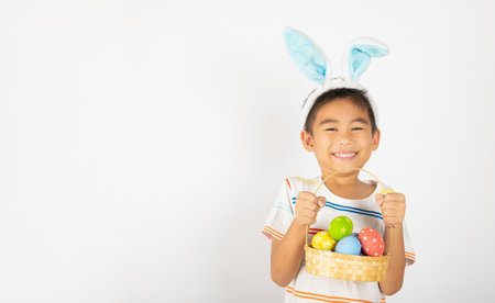 Happy Easter Day. Smile Asian little boy wearing easter bunny ears holding basket of full colorful eggs smiles broadly isolated on white background with copy space, Happy child in holidayの写真素材