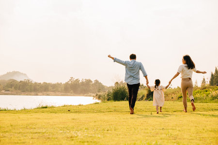 Back of Asian family enjoying a morning walk in a beautiful park, with green fields and a sunny background, smiling and holding hands, Family dayの写真素材