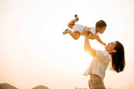 A happy mother holds her baby up in the air in the park, while the child enjoys a moment of playful freedom and joy. Love and family captured in a photographの写真素材