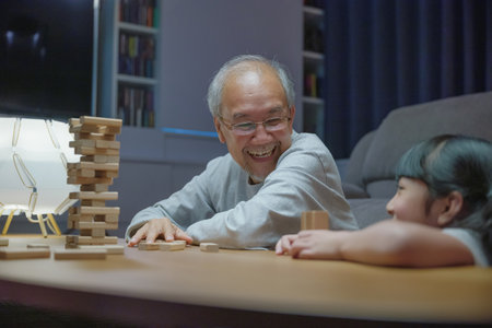 Happy grandparents Asian family enjoy playing toy block with little daughter and mother together at home night time, Smiling parent having fun play build constructor tower of wooden blocks, educationの写真素材