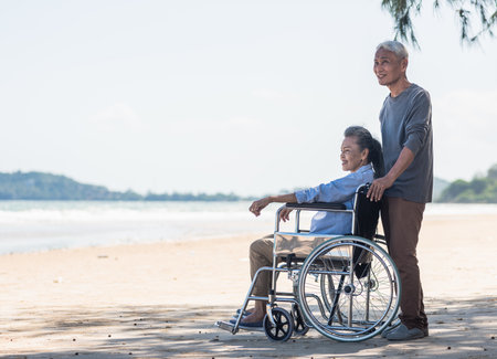 Happy Asian back elderly woman disabled sitting in wheelchair and husband is a wheelchair user on the beach together, summer vacation, Retirement couple conceptの写真素材