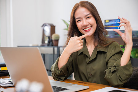 Excited woman holding credit card and pointing the black card with laptop computer at home office, paying shopping online, Beautiful female using laptop while sitting on desk, E-commerceの写真素材