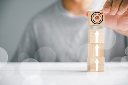 Hand of a businessman arranging wooden blocks in a step-like structure, positioning a cube with a target icon on the highest point. Illustrating the concepts of growth, success, and strategy.の写真素材