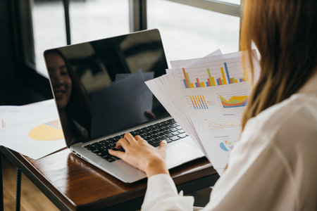 Businesswoman working and comparing forecasting and graphic documents paperwork with laptop computer, Happy female holding papers review report analyzing work results on graph chart outside officeの写真素材