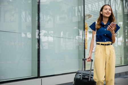 A fashionable young woman walks confidently down a city street with her suitcase in tow, using her mobile phone to stay on top of her busy schedule.の写真素材