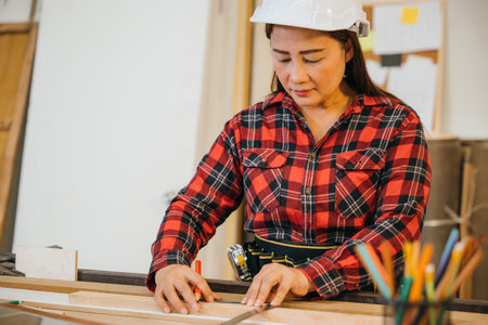 Asian senior woman carpenter taking measurement of a wooden plank and make in workshop, female holding ruler and pencil while making marks on wood at woodshop, Happy carpenter dayの写真素材