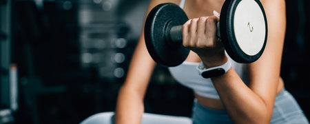A muscular female lifting dumbbells on a bench at the gym to exercise her arm muscles and build up her strength and endurance for an active lifestyle, fitness GYM dark backgroundの写真素材