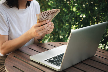 Close up hands of woman holding many money thai baht after she working on laptop computer outside in cafe coffee shop, Achievement career conceptの写真素材