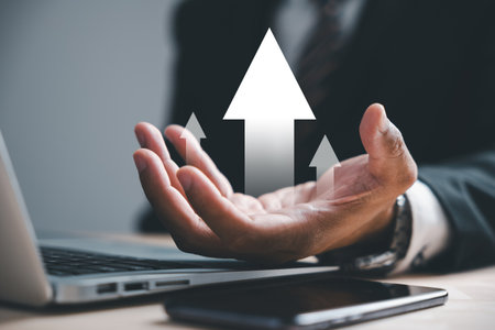 Businessman holds arrow graph on palm with laptop computer, depicting development to success and growing growth concept. Corporate future growth plan in hand illustrates business strategy.の写真素材
