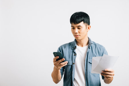 A young man, isolated on white, reads a receipt and answers a mobile phone call while holding bills and paperwork, portraying his financial stress and dedication.の写真素材
