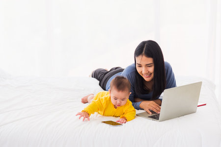 Asian young mother work from home busy on laptop computer and his baby daughter playing smartphone,...