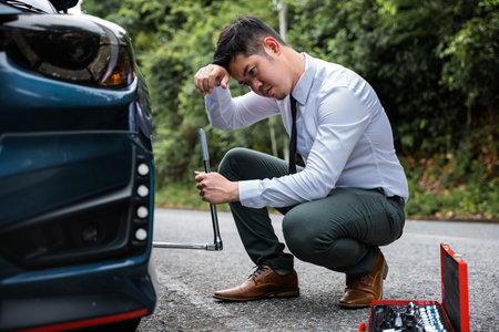 Confident young man in uniform changing car tyre on the roadside. Roadside assistance by Asian businessman wearing white shirt. Expert car mechanic at work.の写真素材