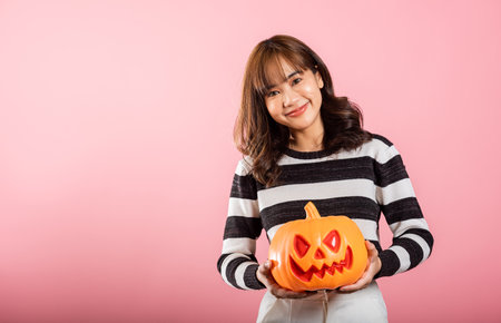 An Asian woman embraces the Halloween spirit in a studio shot on pink background, holding model pumpkins, including one in the shape of a ghost. Her cheerful costume adds to the fun.の写真素材